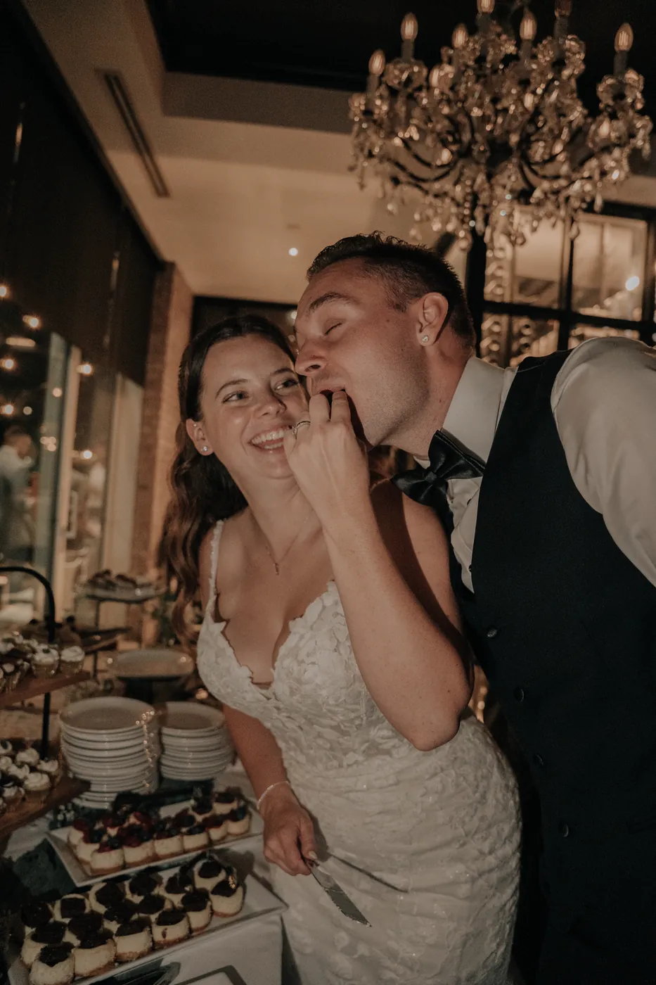 The couple sharing a playful moment beside a dessert table under a chandelier.