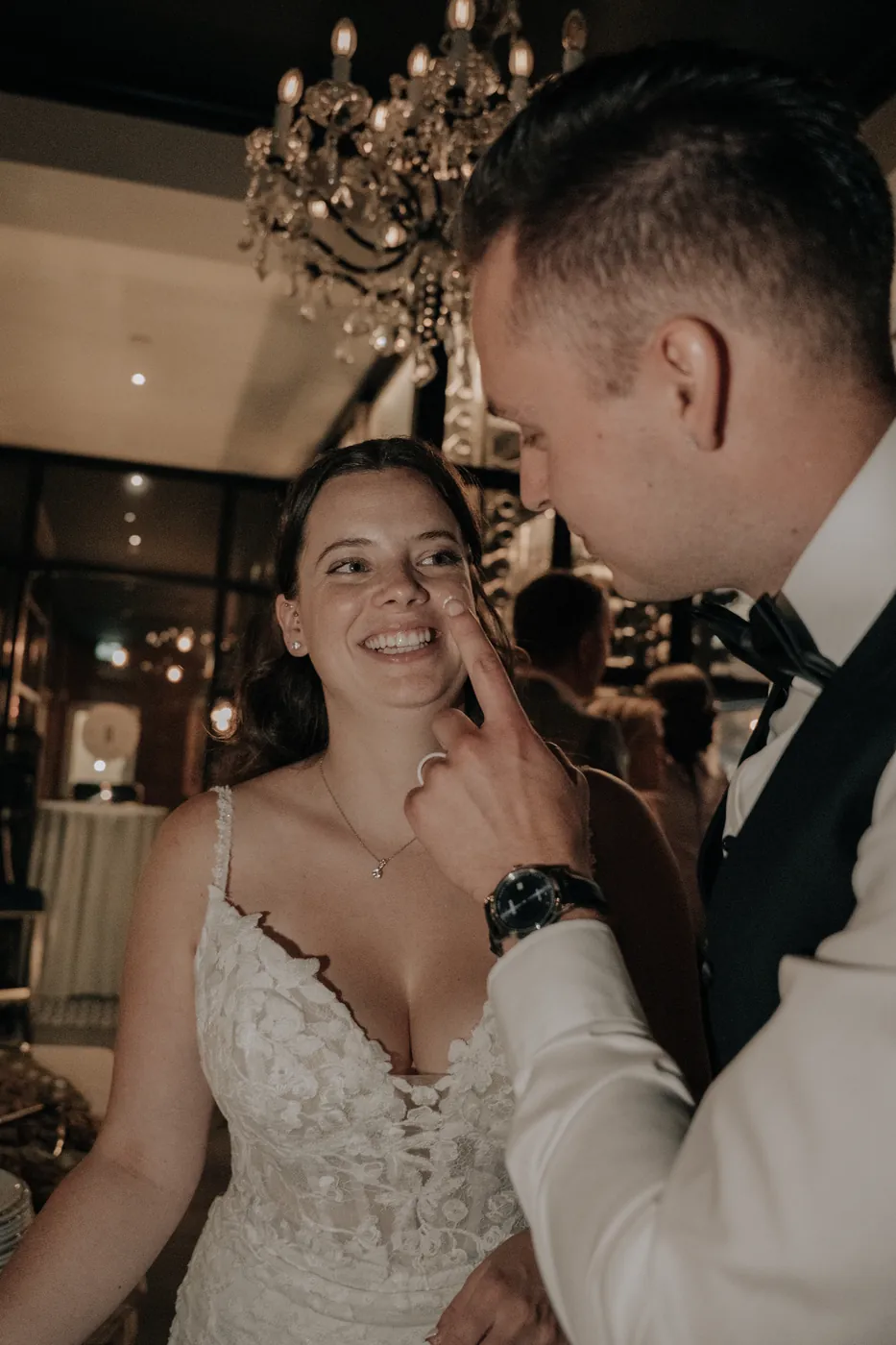 A smiling bride with the groom at a chandelier-lit reception.